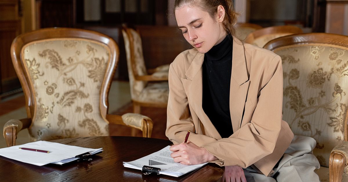 Young woman in a brown blazer signing documents at a table in a stylish office.