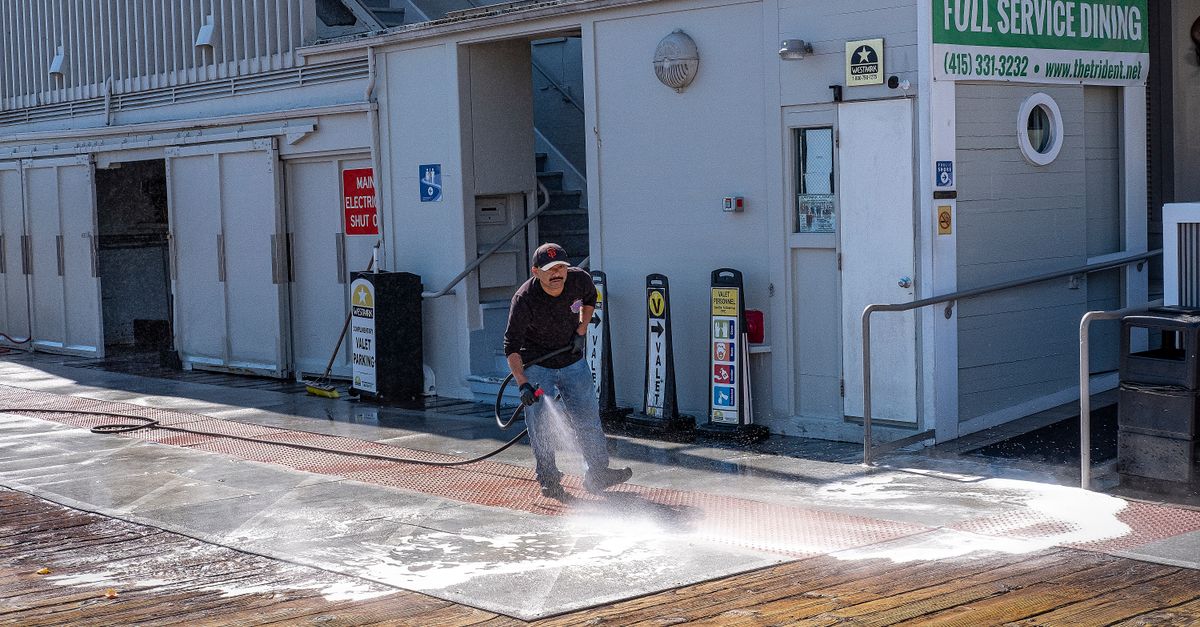 A worker uses a pressure washer to clean a sidewalk outside a dining facility.