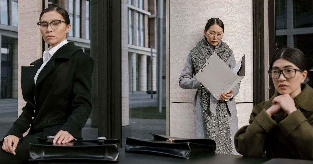 Three professional women in formal outfits with briefcases, posing outside a modern office building.