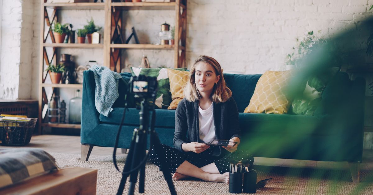 Woman filming vlog at home with camera setup, surrounded by plants and cozy decor.